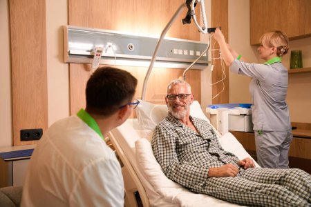 Téléchargez les photos : Le patient en pyjama communique avec le médecin traitant dans un service hospitalier, à côté d'une infirmière qui lui prépare une intraveineuse - en image libre de droit