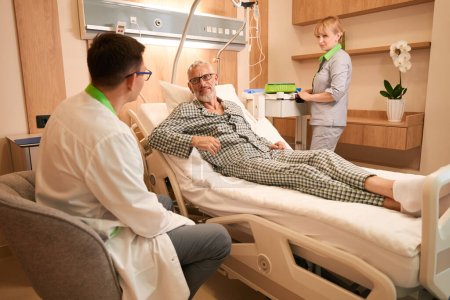 Photo for Elderly man communicates with the attending physician in a hospital ward, next to a nurse preparing an IV for him - Royalty Free Image