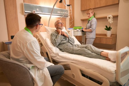 Téléchargez les photos : Le médecin écoute les plaintes d'un patient âgé, une infirmière lui prépare une intraveineuse - en image libre de droit