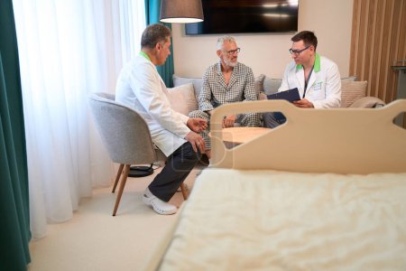 Téléchargez les photos : Les médecins communiquent avec un patient dans une chambre d'hôpital confortable, les gens s'assoient sur un canapé confortable dans l'espace détente - en image libre de droit