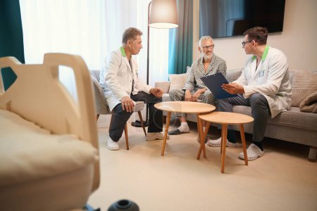 Téléchargez les photos : Des collègues médicaux interrogent un vieil homme dans une chambre confortable, les gens sont assis sur un canapé confortable - en image libre de droit
