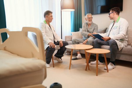 Téléchargez les photos : Des collègues médicaux interrogent un vieil homme dans une chambre confortable, le patient serre la main du médecin traitant - en image libre de droit