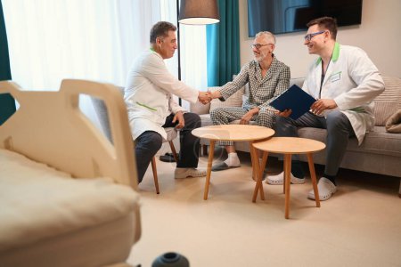 Téléchargez les photos : Vieil homme dans une chambre confortable serre la main du médecin traitant, d'autres thérapeutes conseillent le patient - en image libre de droit