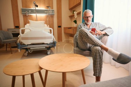 Téléchargez les photos : Homme en pyjama d'hôpital assis avec un téléphone portable sur une chaise douce dans une chambre confortable - en image libre de droit