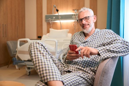 Téléchargez les photos : Vieil homme en pyjama à carreaux se repose après les procédures, il est assis avec son téléphone portable sur une chaise douce - en image libre de droit