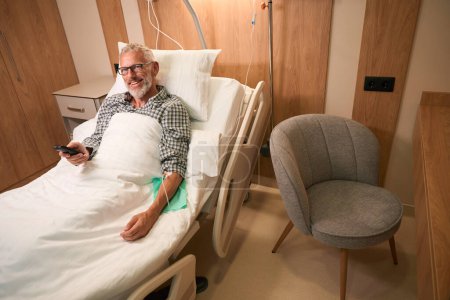 Téléchargez les photos : Homme aux cheveux gris couché sous un goutte à goutte dans une chambre confortable, il subit un traitement dans un établissement médical moderne - en image libre de droit
