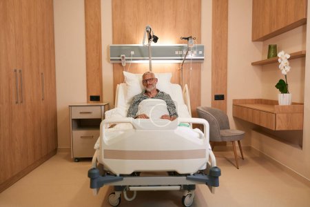 Téléchargez les photos : Vieil homme installé confortablement dans la chambre d'hôpital, la chambre était lumineuse et propre - en image libre de droit