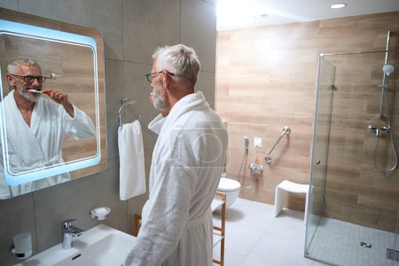 Téléchargez les photos : Vieil homme joyeux se brosse les dents dans la salle de bain, il porte un peignoir éponge moelleux - en image libre de droit