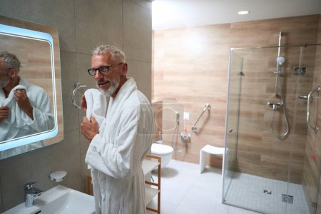 Téléchargez les photos : Vieil homme en peignoir essuie son visage avec une serviette devant un miroir, il a une barbe et des lunettes - en image libre de droit