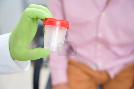 Téléchargez les photos : Un employé de clinique donne à un homme un récipient pour recueillir un test d'urine, un médecin travaille dans des gants de protection - en image libre de droit
