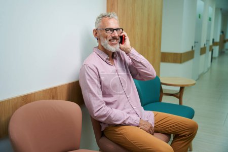 Photo for Old man communicates on his mobile phone in a hospital corridor, the room is bright and clean - Royalty Free Image