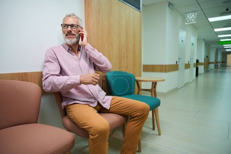 Téléchargez les photos : Homme à lunettes communique sur un téléphone portable dans un couloir d'hôpital, la chambre est lumineuse et propre - en image libre de droit