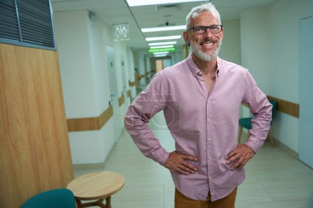 Téléchargez les photos : Homme souriant dans une chemise rose se tient dans un couloir d'hôpital, la chambre est lumineuse et propre - en image libre de droit