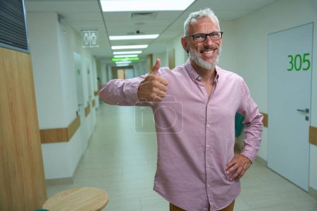 Téléchargez les photos : Un homme souriant en chemise rose montre ok, le couloir de l'hôpital est vide et propre - en image libre de droit