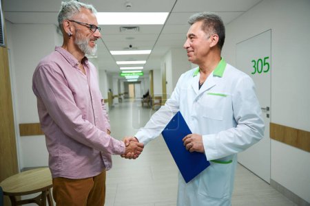 Téléchargez les photos : Le docteur accueille un homme dans un couloir lumineux de l'hôpital, ils se serrent la main - en image libre de droit