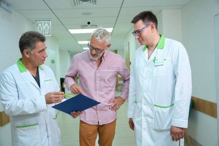Téléchargez les photos : Trois hommes, deux médecins et un patient, marchent le long d'un couloir d'hôpital, ils discutent des papiers d'assurance - en image libre de droit