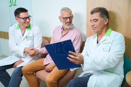 Téléchargez les photos : Les médecins communiquent avec un homme aux cheveux gris dans un couloir d'hôpital, ils s'assoient sur des chaises confortables - en image libre de droit
