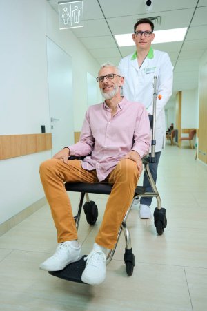 Téléchargez les photos : Employé de la clinique porte un vieil homme en fauteuil roulant le long du couloir de l'hôpital, la chambre est propre et lumineuse - en image libre de droit