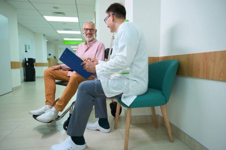 Téléchargez les photos : Médecin traitant en blouse médicale conduit un questionnaire sur un patient, un homme souriant assis dans un fauteuil roulant - en image libre de droit