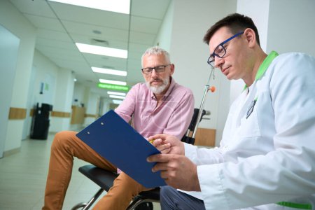Téléchargez les photos : Un homme en fauteuil roulant et un médecin parlent dans un couloir d'hôpital, ils discutent des papiers d'assurance - en image libre de droit