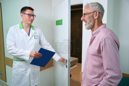 Téléchargez les photos : Docteur a ouvert la porte du cabinet médical, il parle à un homme en chemise rose - en image libre de droit