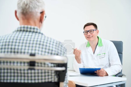 Téléchargez les photos : Homme en blouse médicale communique avec un vieil homme, un patient en pyjama d'hôpital assis dans un fauteuil roulant - en image libre de droit