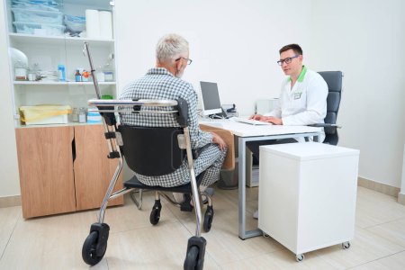 Téléchargez les photos : Thérapeute conduit un rendez-vous dans un cabinet de médecins, un patient en pyjama d'hôpital assis dans un fauteuil roulant - en image libre de droit