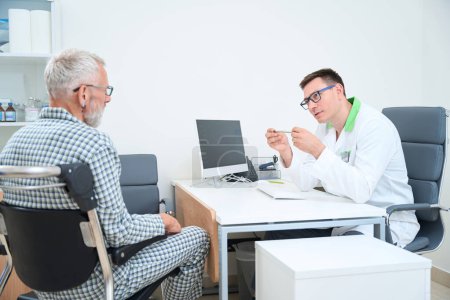 Téléchargez les photos : Thérapeute communique avec un homme aux cheveux gris dans un cabinet de médecins, un patient en pyjama d'hôpital assis dans un fauteuil roulant - en image libre de droit