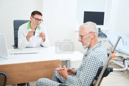Téléchargez les photos : Médecin traitant communique avec l'homme triste dans le bureau des médecins, patient en pyjama d'hôpital se trouve en fauteuil roulant - en image libre de droit