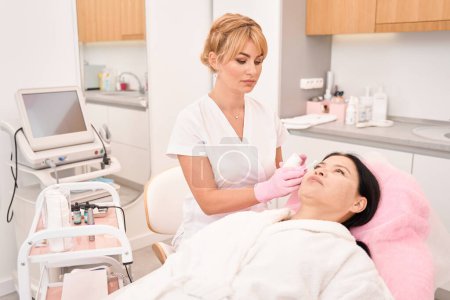 Photo for Esthetician applies cream to a woman face with an applicator, the client sits comfortably in a cosmetology chair - Royalty Free Image