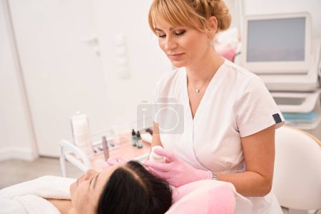 Photo for Beauty master applies cream to a woman face with an applicator, the client sits comfortably in a cosmetology chair - Royalty Free Image