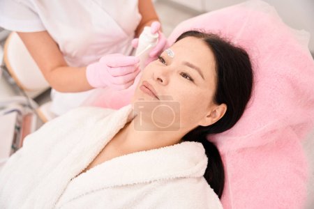 Téléchargez les photos : Beauty master applique de la crème sur les sourcils d'une femme avec un applicateur, le client s'assoit confortablement dans une chaise de cosmétologie - en image libre de droit