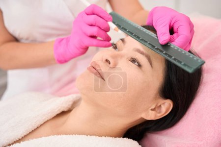 Photo for Esthetician uses a special pencil and ruler to mark the clients eyebrows before the procedure, the master works with gloves - Royalty Free Image