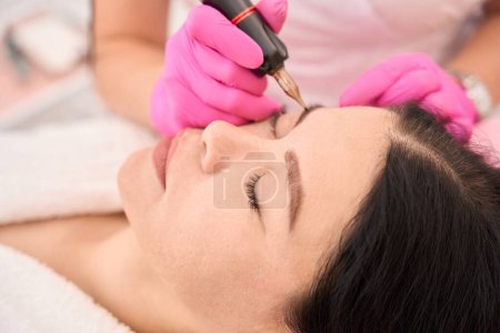 Téléchargez les photos : Femme en gants roses fait un tatouage des sourcils d'un client, pigment de haute qualité est utilisé - en image libre de droit