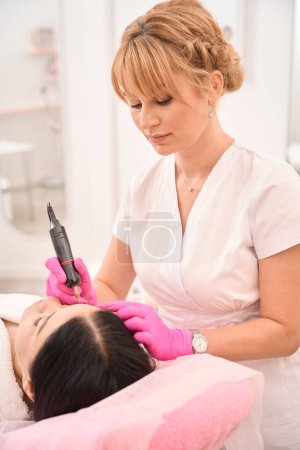 Photo for Beautiful blonde makes a tattoo of eyebrows to a client, the artist works in protective gloves - Royalty Free Image