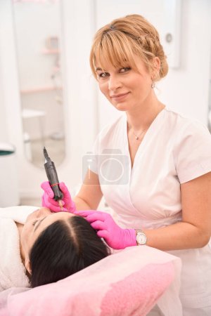 Photo for Charming blonde makes a tattoo of eyebrows to a client, the artist works in protective gloves - Royalty Free Image