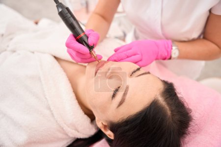 Photo for Specialist applies permanent lip makeup to a client using a special apparatus - Royalty Free Image