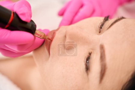 Foto de Belleza artista en guantes de protección hace un tatuaje labial para un cliente, utilizando un aparato especial - Imagen libre de derechos