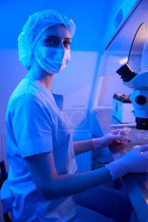 Photo for Calm female scientist sitting on front of microscope with vial filled with liquid solution in her hand - Royalty Free Image