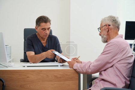 Photo for Gray-haired man signs documents in doctors office, men sit at office desk - Royalty Free Image