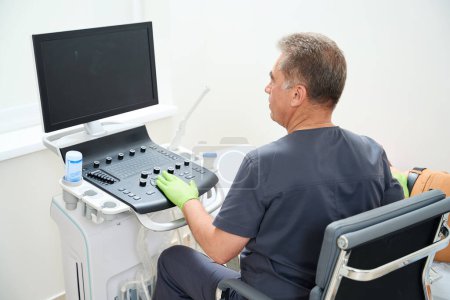 Téléchargez les photos : Médecin sur le lieu de travail effectue un examen échographique, le patient est assis sur le canapé - en image libre de droit