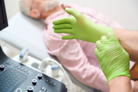 Téléchargez les photos : Spécialiste met des gants de protection avant d'examiner un patient, il y a une machine ultrasonore moderne dans la pièce - en image libre de droit