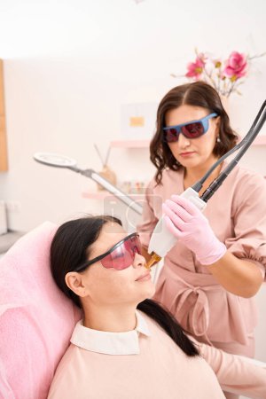 Foto de Procedimiento moderno de depilación facial con láser en una clínica de cosmetología, mujeres con gafas protectoras - Imagen libre de derechos
