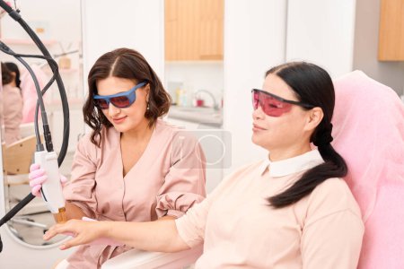 Téléchargez les photos : Client d'une clinique de cosmétologie soumis à une procédure d'épilation au laser des doigts, personnes portant des lunettes de sécurité - en image libre de droit
