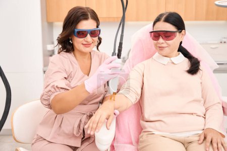 Téléchargez les photos : Femme cosmétologue et cliente de salon dans des lunettes de protection, subissant une procédure d'épilation au laser - en image libre de droit
