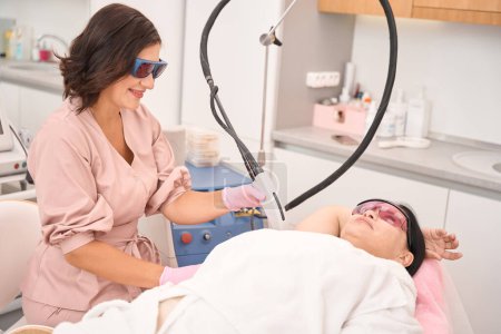 Photo for Beauty master doing laser hair removal on clients armpits, woman wearing safety glasses - Royalty Free Image