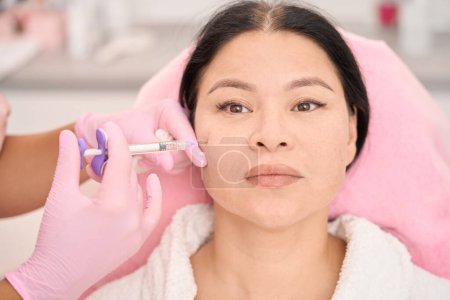 Téléchargez les photos : Belle femme asiatique subissant une procédure de rajeunissement par injection dans un salon de cosmétologie, un spécialiste travaille en gants de protection roses - en image libre de droit