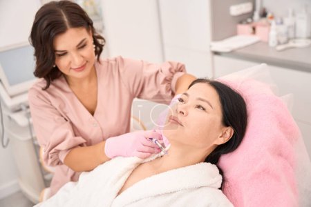 Téléchargez les photos : Femme cosmétologue donne des injections de beauté à la région du cou d'un client asiatique, à l'aide d'une petite seringue - en image libre de droit