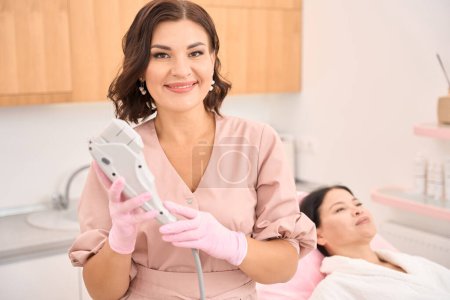 Photo for Female cosmetologist with a device for smas lifting, next to her in a cosmetology chair is a smiling client - Royalty Free Image