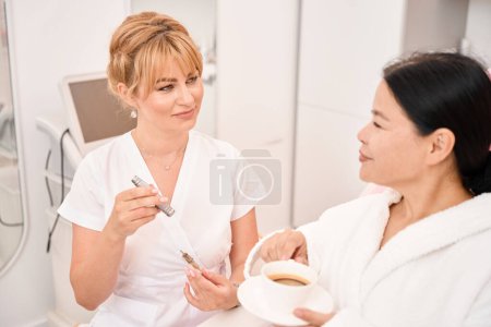 Foto de Cliente habla sobre una taza de café con un artista de maquillaje permanente, el especialista tiene un dispositivo especial - Imagen libre de derechos
